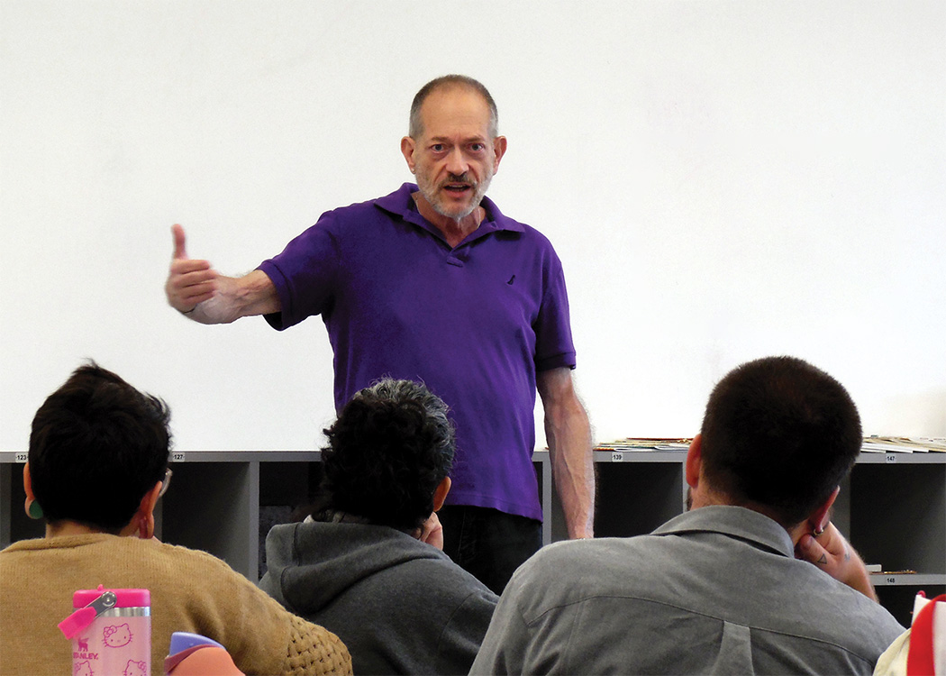 Man with short hair wearing purple short-sleeve polo shirt stands and lectures with his right arm extended mid-sentence in front of a class of seated students