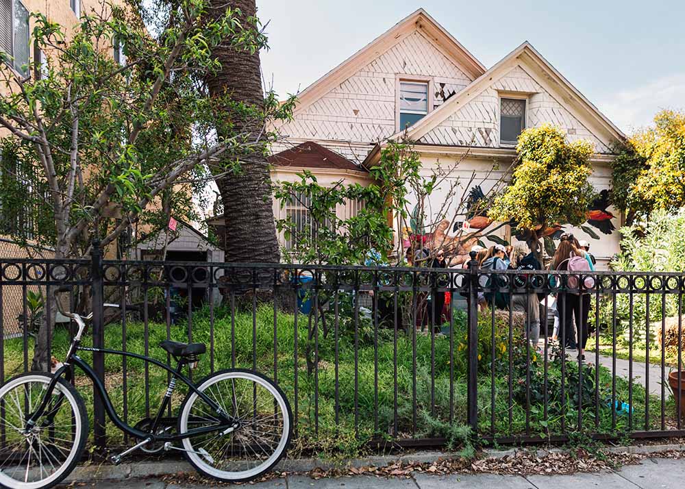 People gather in an urban garden setting as seen from the street with bicycle and low gate in the foreground