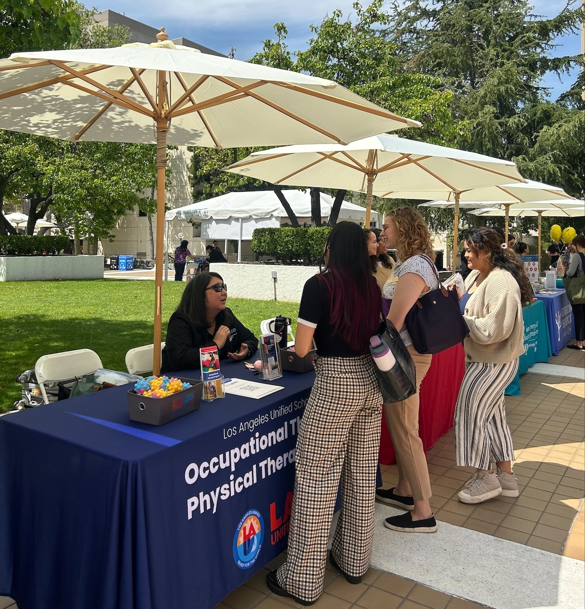 Several people with backpacks stand in front of a table set up in an outdoor courtyard with white umbrellas. The tablecloth says Occupational THerapy and Physical Therapy