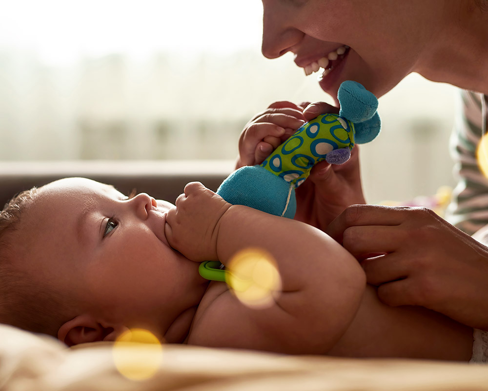 Close-up photo of infant lying on back with toy in mouth and gazing at mother