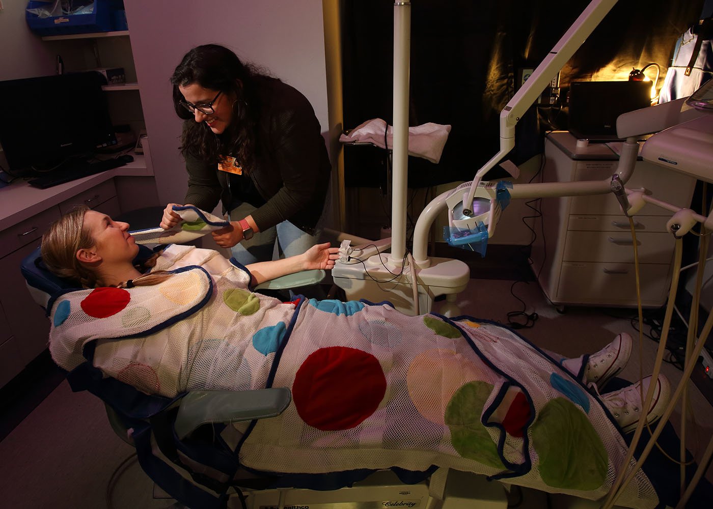 Photograph of a young person reclined in a dental operatory chair while a researcher secures a mesh style wrap around the chair.