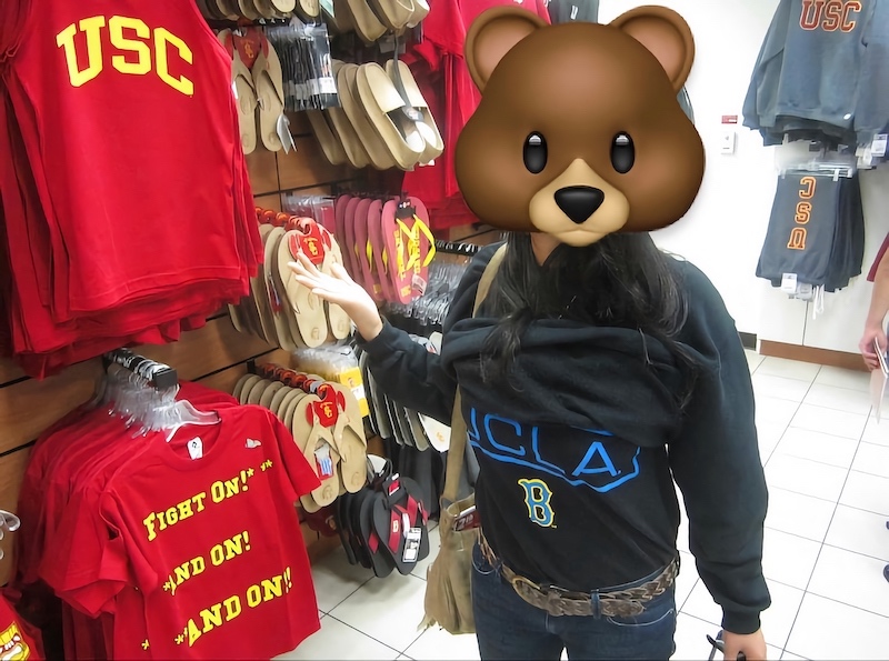 A photo of Teresa in May 2012 standing in the USC Bookstore. She has superimposed a bear emoji on top of her face. She is wearing a UCLA shirt and gesturing to the USC shirts hanging in the store.